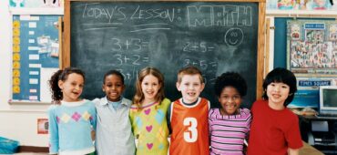 Kindergarten students of different races stand in front of classroom blackboard